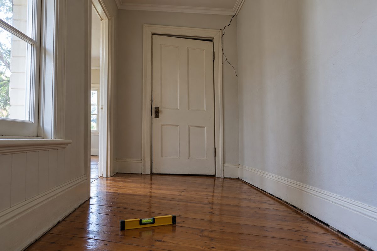 Inside an older weatherboard home in Warragul, a hallway of polished baltic pine floorboards visibly dipping toward one wall, a painted timber door hanging out of square with an uneven gap along the top, a diagonal crack in the plaster from the door frame to the cornice, a gap under the skirting and a spirit level on the floor with its bubble off centre